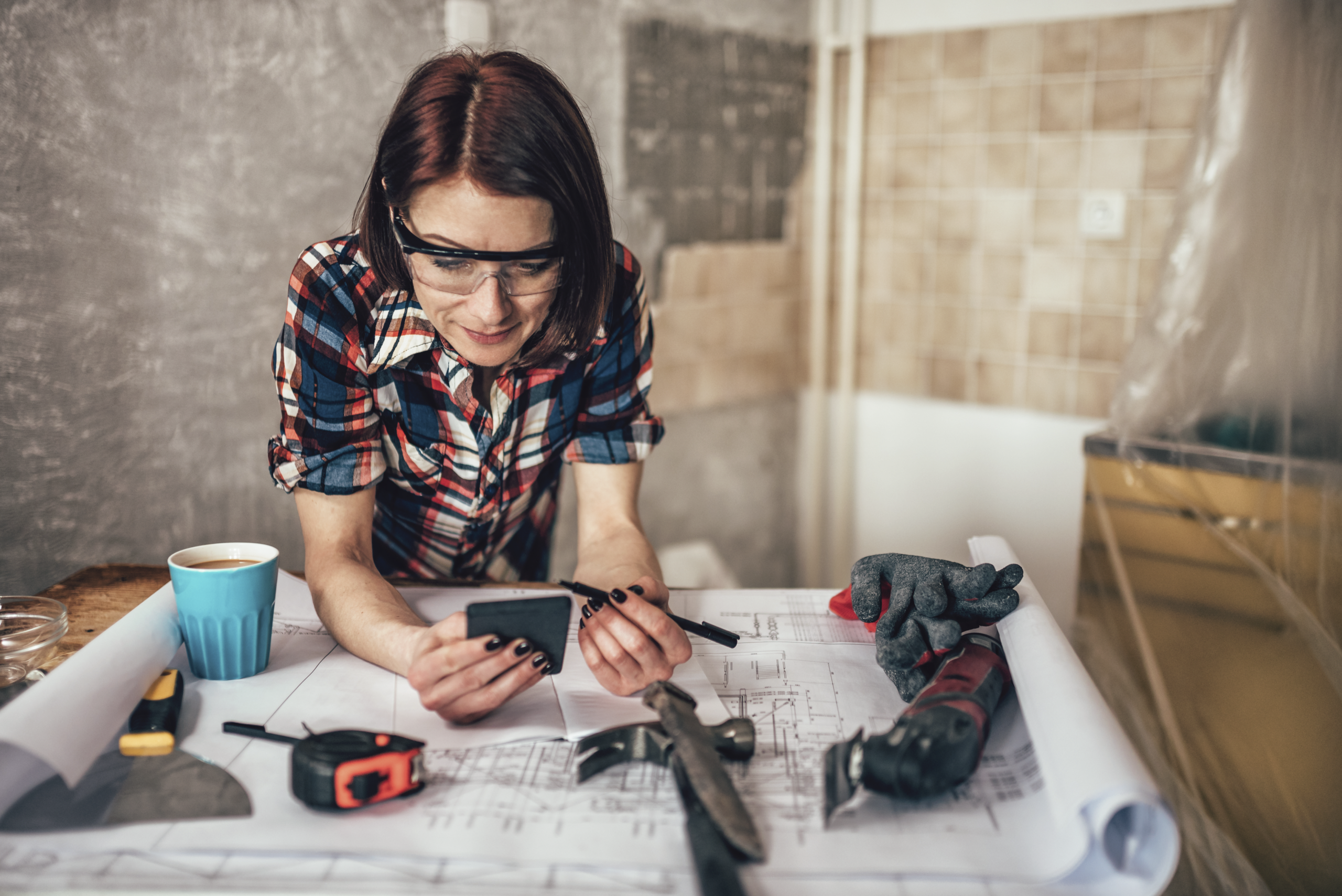 DIY homeowner using a renovation cost estimator on a phone while reviewing blueprints for a specific project.